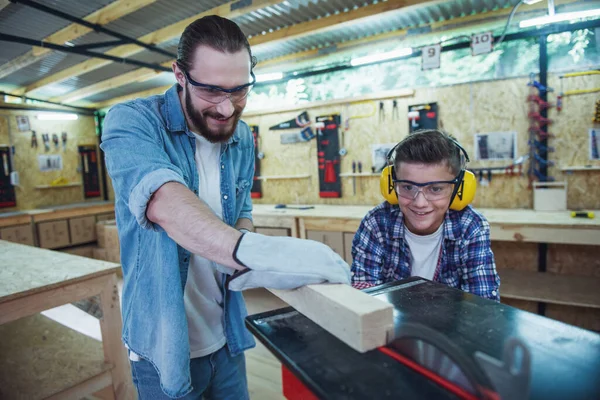 Handsome teenage carpenter and his father in protective glasses are smiling while working with wood and a circular saw table in the workshop