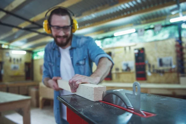 Handsome carpenter in protective glasses and headphones is smiling while working with wood and a circular saw table in the workshop