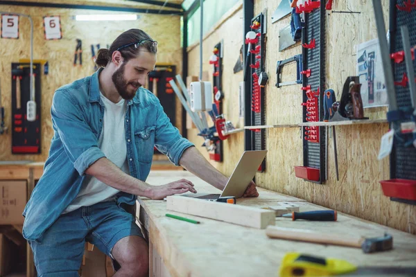 Handsome carpenter is smiling and using a laptop while working in the workshop