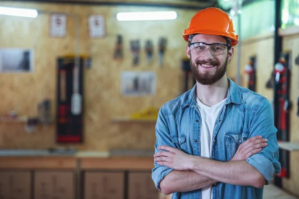Handsome carpenter in hard hat and protective glasses is looking at camera and smiling while standing with crossed arms in the workshop