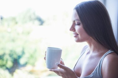 Bir fincan holding, kahve kokulu ve pencerenin evde yakınındaki Kapalı gözlerle otururken gülümseyen gri singlet güzel seksi genç kadını yan görünüm