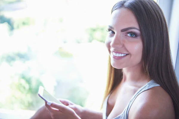 美丽性感的年轻女子 在灰色的单线态是使用智能手机 微笑着坐在家里的窗台上 — 图库照片
