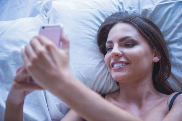 美丽性感的年轻女子 在黑色的胸罩 使用智能手机和躺在床上微笑着的肖像 — 图库照片