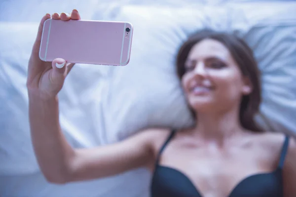 美丽性感的年轻女子 在黑色的胸罩 使用智能手机和躺在床上微笑着的肖像 — 图库照片