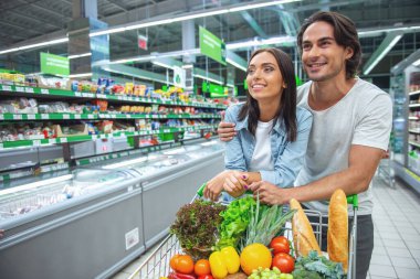 Güzel genç çift üzerinde alışveriş sepeti yaslanmış, kameraya bakıyor ve süpermarket gıda seçerken gülümseyen