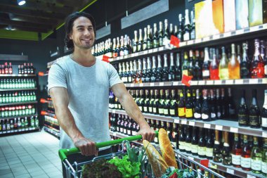 Yakışıklı genç bir alışveriş sepeti iterek ve süpermarkette içecekler seçerken gülümseyen
