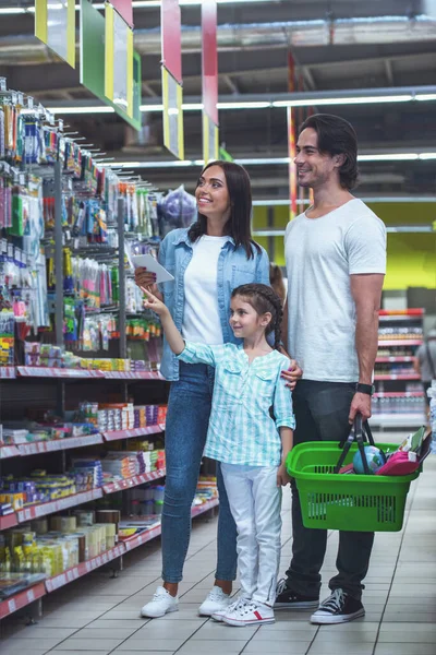 Beautiful young parents and their cute little daughter are smiling while choosing school stationery in the supermarket. Girl is pointing