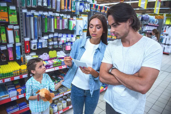 Beautiful young parents and their cute little daughter are choosing school stationery in the supermarket. Mom and dad are discussing the list