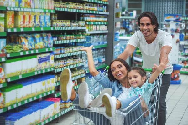 Cheerful young family, mom and dad and their cute little daughter pushing a shopping cart, looking at the camera and smiling in a supermarket