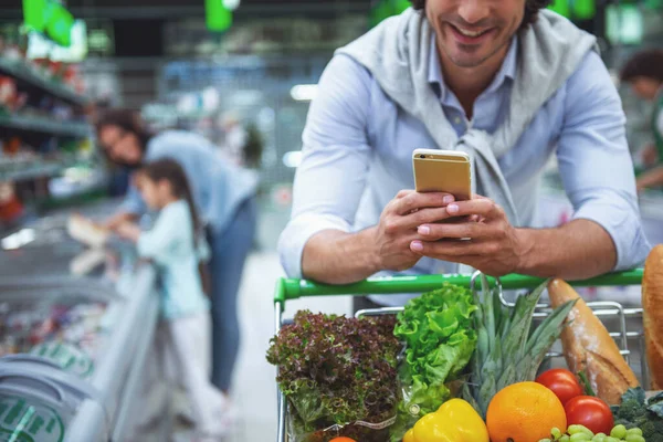 Family in the supermarket. Cropped image of dad leaning on shopping cart, using a mobile phone and smiling, in the background his wife and daughter are choosing food