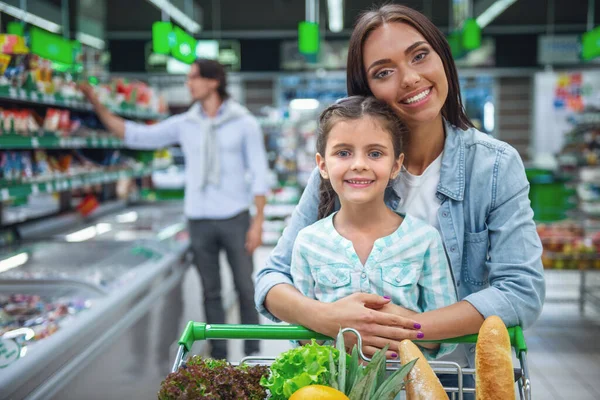 Family in the supermarket. Beautiful young mom and her little daughter are looking at camera and smiling, in the background dad is choosing goods
