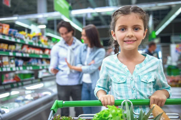 Family in the supermarket. Cute little girl is looking at camera and smiling, in the background her parents are choosing goods