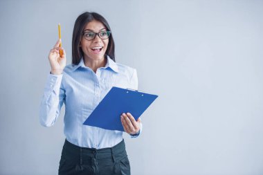 Akıllı rahat giyim ve gözlük güzel genç kız gri arka plan üzerinde, uzağa bakarak ve gülümseyerek, bir klasör ve kalem tutuyor