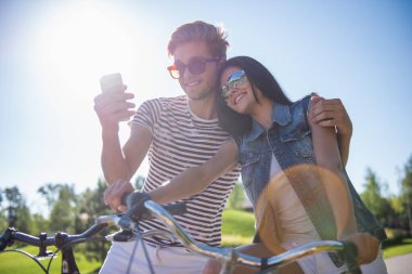 Güzel çift bir smartphone kullanarak ve parkta Bisiklete binme süre gülümseyen selfie yapıyor