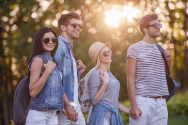 İki güzel şık çift sırt çantaları ile el ele tutuşarak ve açık havada dinlenme süre gülümseyerek. Bir kız kameraya bakıyor