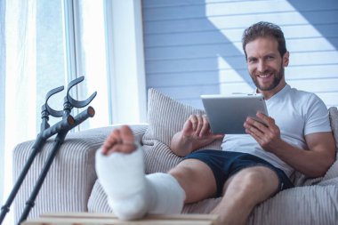 Yakışıklı adam kırık bacak ile dijital tablet kullanma, kameraya bakıyor ve evde kanepede otururken gülümseyen