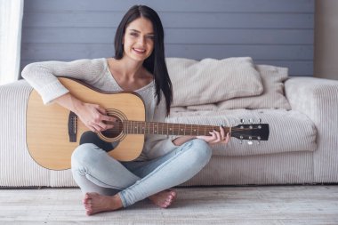 Çekici genç kız gitar çalmak, kameraya bakıyor ve yerde evde otururken gülümseyen