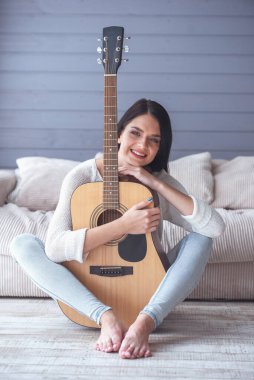Çekici genç kız bir gitar holding, kameraya bakıyor ve yerde evde otururken gülümseyen