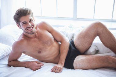 Siyah külot seksi genç adam kameraya bakıyor ve yatakta yatan gülümseyen