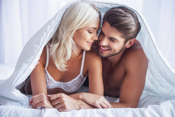 Stock image Beautiful young couple is hugging and smiling while lying covered with blanket in bed