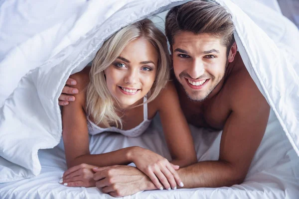 Güzel genç çift sarılma, kameraya bakıyor ve yatakta battaniye ile örtülü yatan gülümseyen