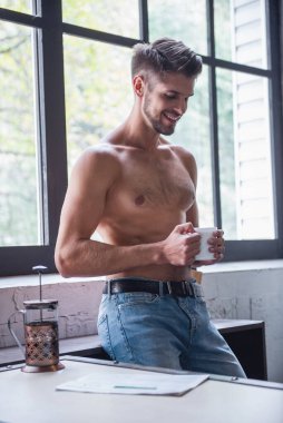 Seksi genç adam çıplak gövde ile bir fincan tutarak ve evde mutfakta dururken gülümseyen