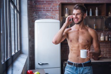 Seksi genç adam çıplak gövde ile bir fincan holding, cep telefonda konuşurken ve evde mutfakta dururken gülümseyen