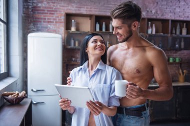 Güzel genç çift dijital tablet kullanma, birbirine bakarak ve evde mutfakta dururken gülümseyerek. Adam bir bardağı götürüyor