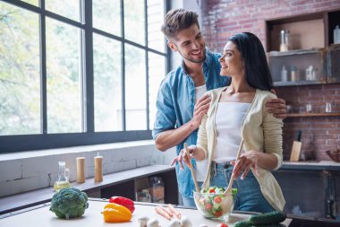 Güzel genç çift birbirine bakarak ve evde mutfakta yemek yaparken gülümseyerek. Kadın salata karıştırma
