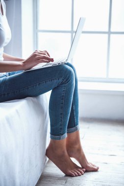 Yatakta evde pencere yanında otururken bir dizüstü bilgisayar kullanarak kot pantolon güzel kızın Resim kırpılmış