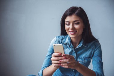 Jean giysili güzel kızı bir smartphone kullanarak ve gri arka plan üzerinde gülümseyen