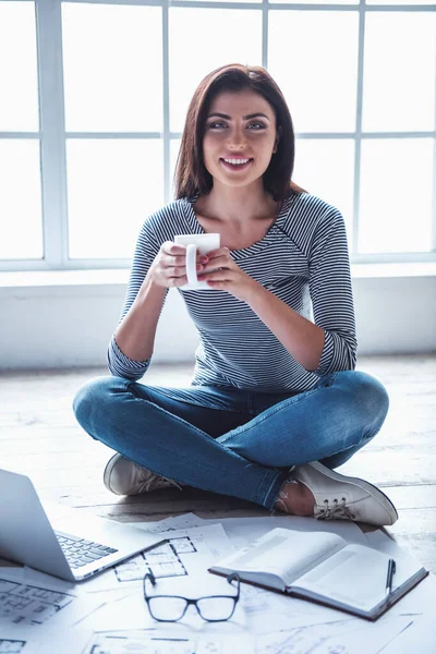Beautiful young businesswoman in casual clothes is holding a cup and smiling while sitting on the floor at home and working