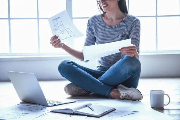 Cropped image of beautiful young businesswoman in casual clothes is studying drafts while sitting on the floor at home and working