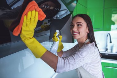 Güzel genç kadın bir deterjan ve bir toz bezi kullanarak ve mutfak temizlerken gülümseyen