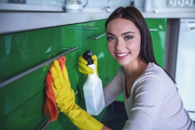 Güzel genç kadın bir deterjan ve bir toz bezi kullanarak, kameraya bakıyor ve mutfak temizlerken gülümseyen