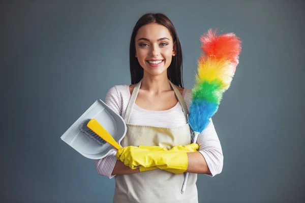 Beautiful young woman is holding a static duster and a dustpan, looking at camera and smiling, on gray background