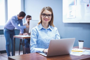 Güzel iş adamları ofisinde çalışıyor. İş kadını kameraya bakıyor ve gülümsüyor, bir dizüstü bilgisayar kullanarak