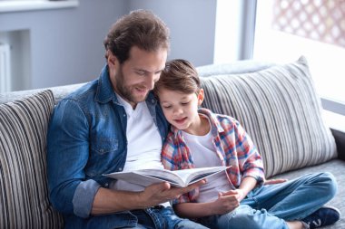 Baba ve oğul evde birlikte vakit geçirirken kitap okuyor ve gülümsüyorlar.