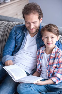 Baba ve oğul evde birlikte vakit geçirirken kitap okuyor ve gülümsüyorlar.