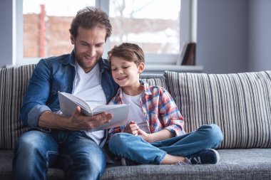 Baba ve oğul evde birlikte vakit geçirirken kitap okuyor ve gülümsüyorlar.