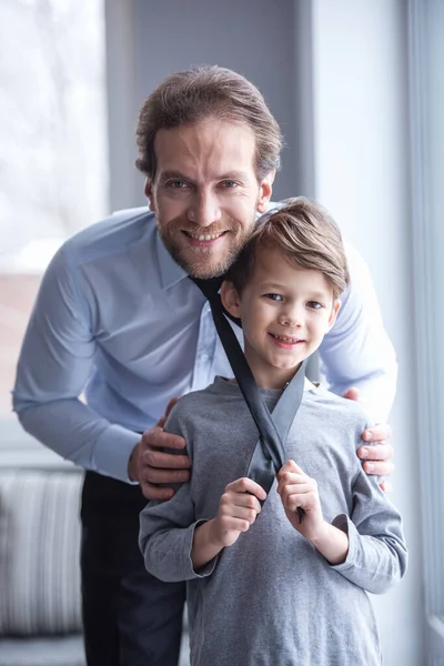Father and son are looking at camera and smiling while spending time together. Little boy is posing with dad\'s tie