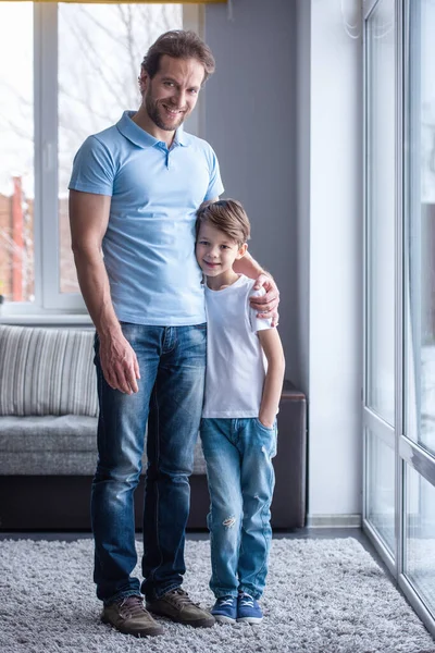 Full length portrait of father and son hugging, looking at camera and smiling while standing in the living room