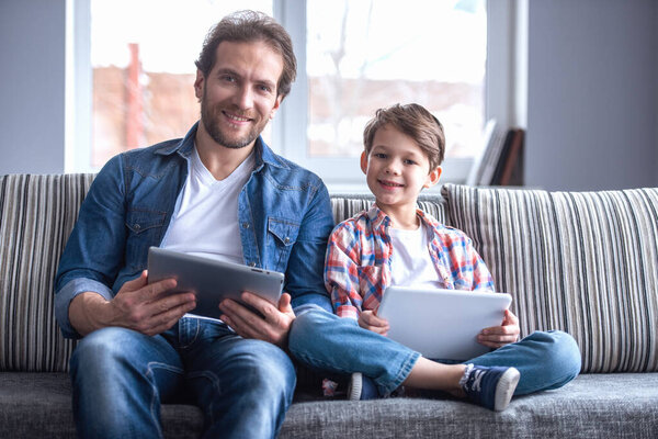 Father and son are using digital tablets and smiling while spending time together at home