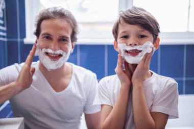 Baba ve oğul traş köpüğü yüzlerinde uygulamak ve banyoda tıraş ederken gülümseyen