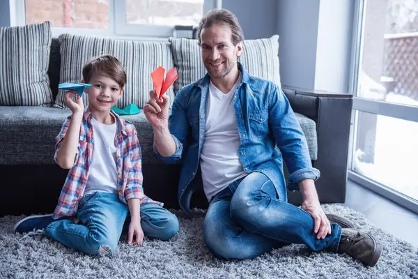 Father and son are playing with paper planes and smiling while spending time together at home