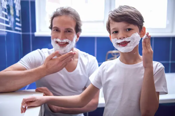 Father and son are applying shaving foam on their faces and smiling while shaving in bathroom
