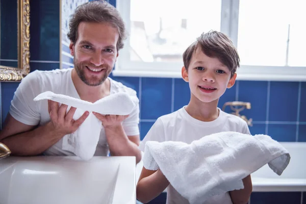 Father and son are holding towels, looking at camera and smiling while washing in bathroom