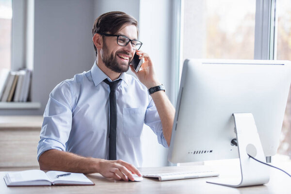 Handsome businessman in eyeglasses is using a computer, talking on the mobile phone and smiling while working in office
