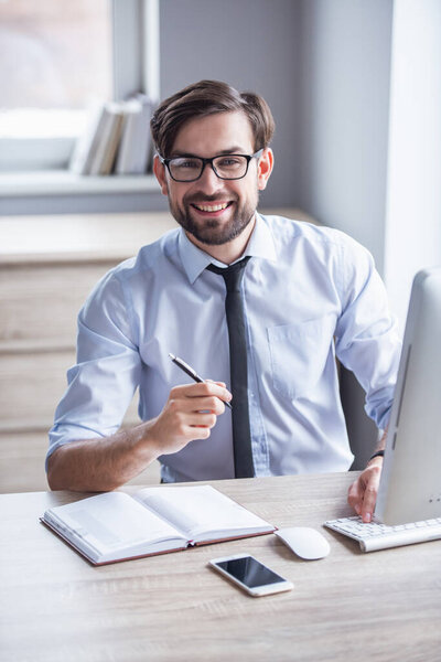 Handsome businessman in eyeglasses is making notes, using a computer and smiling while working in office