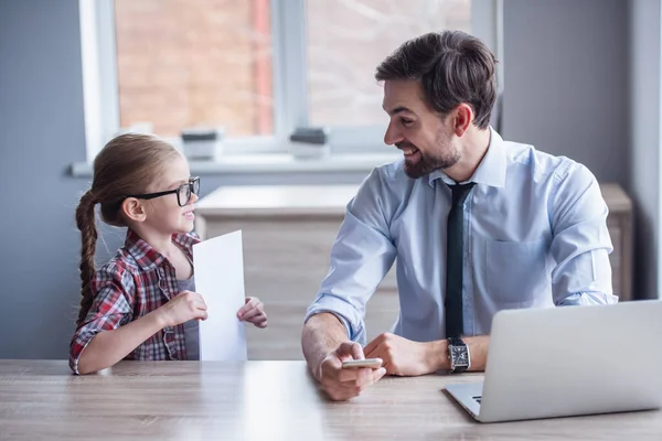 stock image Handsome businessman is using a laptop while working at home. His cute little daughter in glasses is showing him a sheet of paper